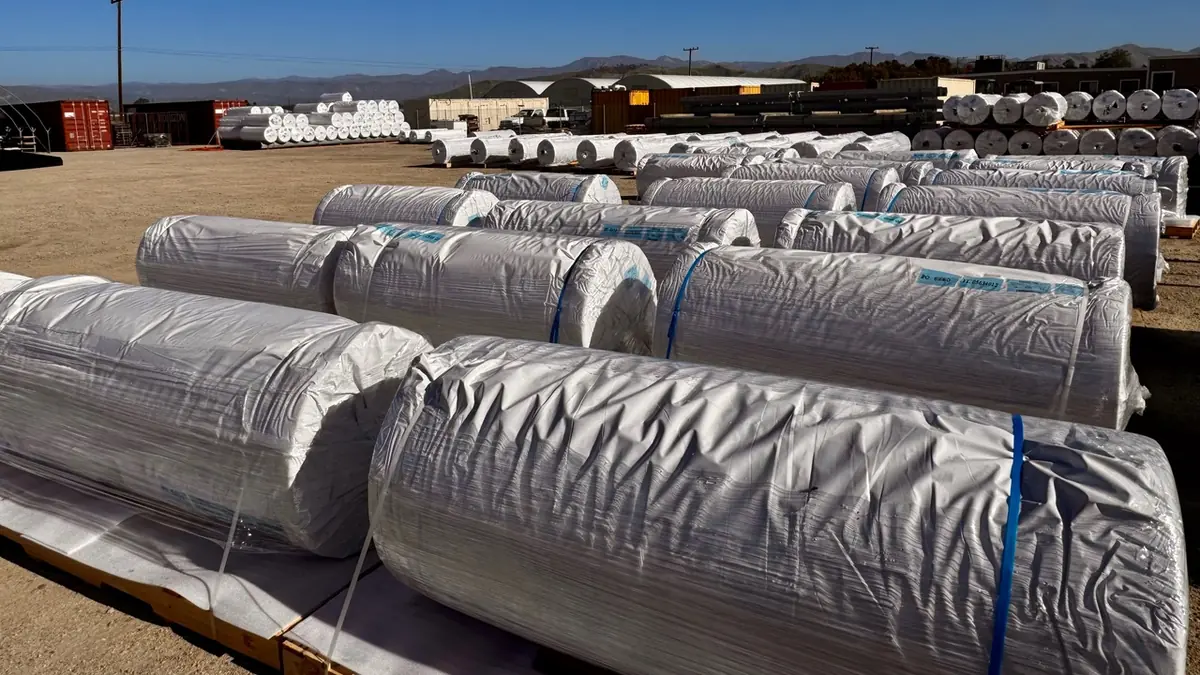 Large wrapped greenhouse film master rolls arranged in rows on wooden supports outdoors.